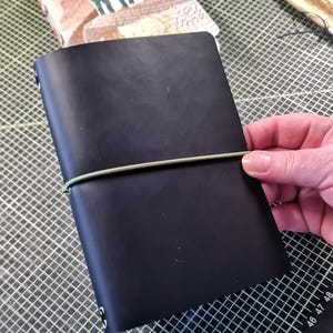 Könnte beinhalten: Ein schwarzes, ledergebundenes Notizbuch mit einem grünen Gummibandverschluss. Das Notizbuch liegt auf einer grün-weißen, karierten Oberfläche.