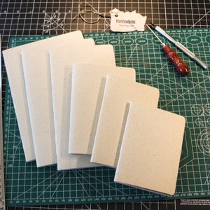 Könnte beinhalten: Eine Sammlung leerer Notizbücher in verschiedenen Größen mit hellen Umschlägen, angeordnet auf einer grünen Schneidematte. Ein Metalllineal und ein Lederwerkzeug sind ebenfalls zu sehen. Das Etikett trägt die Aufschrift "Rainbowbook handmade".