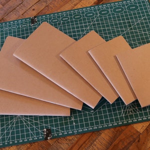 Könnte beinhalten: Fünf braune Notizbücher in verschiedenen Größen, die auf einer grünen Schneidematte übereinander gestapelt sind. Die Matte hat weiße Gitterlinien und ist mit "A3 450 x 300 mm" beschriftet.