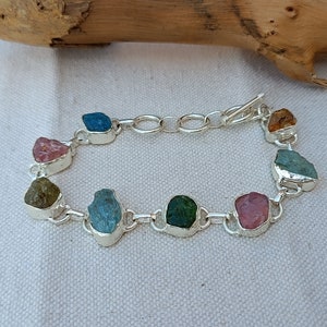 May include: A silver bracelet with a chain link design features eight different colored gemstones. The gemstones are set in silver and are in a variety of shapes and sizes. The bracelet has a lobster claw clasp.