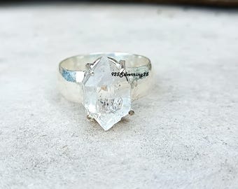 May include: A silver ring featuring a large, clear quartz crystal. The crystal is faceted and has a pointed shape. The ring band is wide and has a hammered texture. The text "925Silverring25" is visible on the ring.