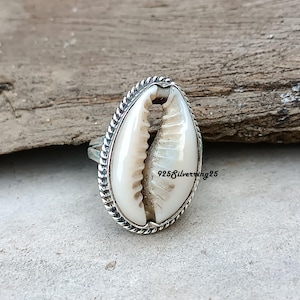 Op de afbeelding: Een zilveren ring met een natuurlijke kaurischelp. De schelp is crèmekleurig met een textuuroppervlak, gezet in een zilveren band met een touwachtige rand. De tekst "925silverring25" is zichtbaar.