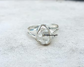 May include: A silver ring with a large, irregularly shaped, clear gemstone. The band splits into two strands on either side of the stone. The text "925 Silverring25" is visible on the gemstone.