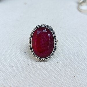 Puede incluir: Anillo ovalado con una piedra de rubí roja. El anillo tiene un borde plateado con forma de cuerda trenzada. El rubí es facetado. El anillo está sobre una superficie blanca. El texto "ShreeJaipurSilver23" es visible.