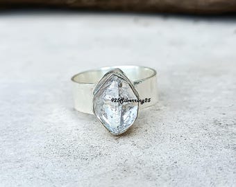 May include: A wide silver ring with a large, clear, faceted gemstone. The gemstone is diamond-shaped and has internal inclusions. The ring band is thick and plain. The text "925silverring25" is visible on the gemstone.