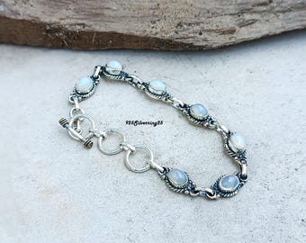 Op de afbeelding: Een zilveren armband met ovale, lichtgekleurde edelstenen. De armband heeft een tuimelsluiting en ligt op een getextureerd oppervlak. De tekst "925Silverring25" is zichtbaar.
