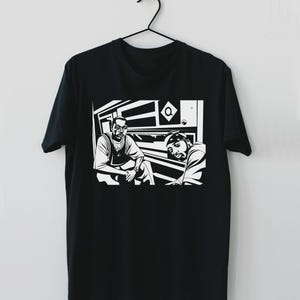 May include: Black t-shirt with a monochrome graphic of two men in a subway carriage. The illustration has a comic book style with strong lines and shading. The t-shirt is suspended from a black hanger.