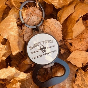 Op de afbeelding: Een zwarte en zilveren flesopener sleutelhanger met de tekst "SLEEPY PINES PREMIUM PROVISIONS" op een witte cirkelvormige achtergrond. De sleutelhanger is geplaatst tegen een achtergrond van gedroogde, bruine herfstbladeren.