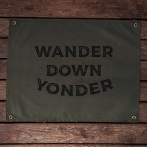 May include: An olive green canvas banner with the text "WANDER DOWN YONDER" printed in black. Metal eyelets are set in each corner. The banner is displayed against a wooden background.