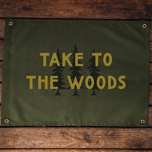 May include: An olive green banner with the words "TAKE TO THE WOODS" in gold. The banner has three silhouetted pine trees. It is mounted on a rustic wooden background.