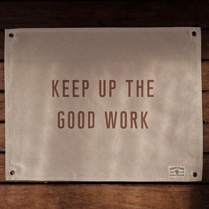 May include: A rectangular, off-white canvas banner with the words "KEEP UP THE GOOD WORK" in a reddish-brown font. The banner has metal grommets in each corner and a small logo in the bottom right corner. The banner is hanging on a wooden wall.