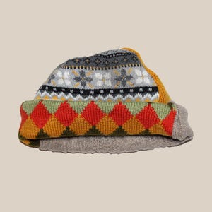 Op de afbeelding: Een gevouwen gebreide beanie-muts met een kleurrijk geometrisch patroon. De muts heeft een grijze basis met een omgeslagen rand met een patroon van rode, oranje en gele ruiten. Het bovenste gedeelte heeft een grijs en wit sneeuwvlokontwerp met gele accenten.