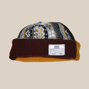 Op de afbeelding: Een gebreide muts met een omgeslagen rand. De bovenkant van de muts heeft een geometrisch patroon in grijs, zwart en geel. De rand is diep bordeauxrood en de omgeslagen rand is mosterdgeel. Een wit label met zwarte tekst is op de rand genaaid.
