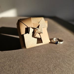 Puede incluir: Un anillo de plata con una piedra preciosa azul oscuro junto a una pequeña caja de regalo cuadrada de estilo origami hecha de papel marrón. La caja está intrincadamente doblada con un diseño en forma de estrella. Sobre una superficie marrón texturizada.