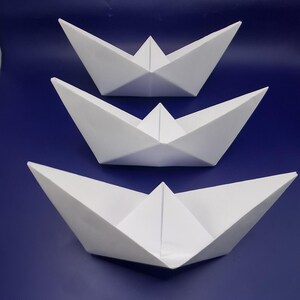 Op de afbeelding: Drie witte papieren bootjes van verschillende formaten op een blauwe achtergrond. De bootjes zijn gemaakt van gevouwen papier, met driehoekige zeilen en open rompen. Het ontwerp is eenvoudig en minimalistisch.