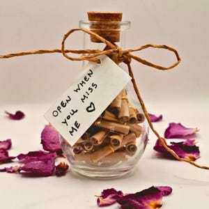 Könnte beinhalten: Eine klare Glasflasche mit Korkverschluss, gefüllt mit kleinen, gerollten braunen Papierrollen. Ein weißes, rechteckiges Etikett, das mit brauner Schnur befestigt ist, trägt die Aufschrift "OPEN WHEN YOU MISS ME" und ein Herzsymbol.