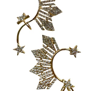May include: A pair of gold ear cuffs with a star design. Each cuff features a star-shaped design with a row of sparkling rhinestones. The cuffs are designed to wrap around the ear and have a curved shape.