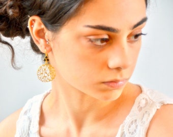 May include: Gold dangle earrings with a circular design featuring intricate filigree work and black beads. The earrings are worn by a person with dark hair.