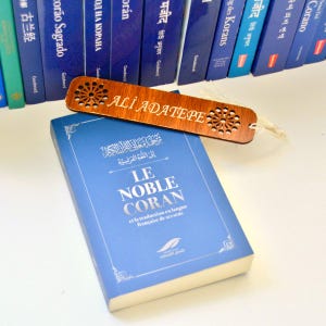 May include: A rectangular wooden bookmark with laser-cut geometric patterns at both ends and the name ALI ADATEPE engraved in the centre, featuring a white tassel, resting on a blue book titled LE NOBLE CORAN with a row of books in the background.