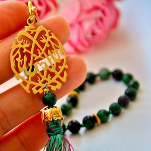 May include: A gold-coloured pendant with intricate Arabic calligraphy is held in a hand. The pendant is attached to a green and gold tassel and a strand of green and black beads. A pink flower is in the background.