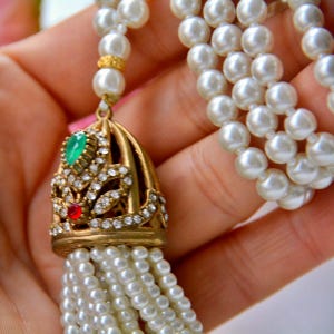 Op de afbeelding: Een vintage parelketting met een gouden hanger. De hanger heeft een smaragdgroene steen, een rode steen en heldere kristallen. De ketting heeft meerdere strengen witte parels en een gouden sluiting.