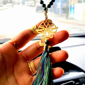 May include: Gold car charm with a tassel. The charm features intricate calligraphy and is attached to a green beaded chain.