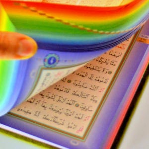 May include: A close-up of a book with pages being turned. The pages have Arabic text. The edges of the pages are rainbow-colored. The book is open, revealing the text and design.