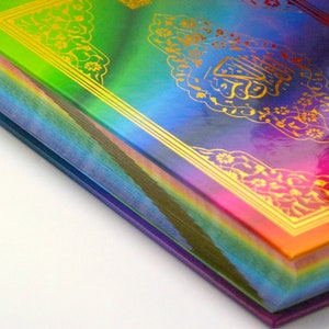 Op de afbeelding: Een hardcover boek met een regenboogkleurige omslag en vergulde Arabische kalligrafie. De randen van de pagina's zijn zichtbaar en tonen een spectrum aan kleuren. De omslag heeft sierlijke gouden bloemmotieven en kalligrafie, wat een levendige uitstraling creëert.