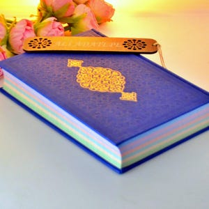 Puede incluir: Un libro azul con un diseño dorado en la portada y un marcapáginas de madera con el texto "Ali Adattepe". El libro está rodeado de flores rosas.