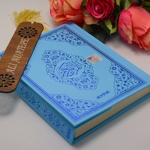Puede incluir: Un libro azul claro con diseños azules ornamentados y el texto "AYFA" se combina con un marcapáginas de madera. El marcapáginas tiene el texto "ALI ADATEPE" y recortes decorativos. Rosas rosas y naranjas en el fondo.