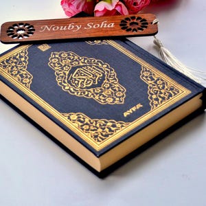 Puede incluir: Un libro azul marino con diseños dorados ornamentados y la palabra "AYFA" en letras doradas. Un marcapáginas de madera con el nombre "Nouby Soha" está colocado encima. El marcapáginas tiene un diseño decorativo recortado.