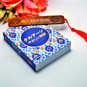 Puede incluir: Un marcapáginas de madera con el nombre "ALI ADATEPE" grabado con láser, colocado sobre un libro con un diseño floral azul y blanco y texto japonés. Una borla roja está unida al marcapáginas. Un ramo de flores naranjas y rosas está en el fondo.