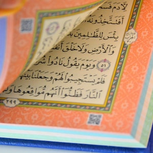 May include: A close-up of an open book with Arabic text. The pages are cream-coloured with black text and a decorative border. The book is bound in a blue cover.