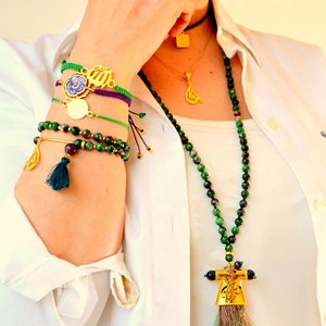 May include: An assortment of gold-toned and beaded jewelry. A long necklace with green and black beads and a gold pendant hangs over a white shirt. Several bracelets with gold charms and green, purple, and gold accents adorn the wrist.