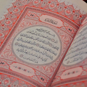 以下が含まれることがあります： アラビア語の筆記体で書かれた開いた本。ページはクリーム色で、赤い装飾的なボーダーとテキストがあります。テキストは黒インクで、上部に「Surat Al-Baqarah」というタイトルがあります。この本は宗教的なテキストのようです。