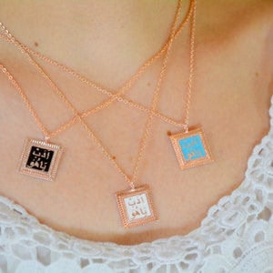 May include: Three rose gold necklaces with square pendants. Each pendant has a different colored background: black, white, and turquoise. The pendants feature Arabic script and are framed with a textured border. The necklaces are displayed on a person's chest.