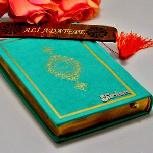Könnte beinhalten: Ein türkisfarbenes Buch mit goldenem Rand und einem goldgeprägten Design. Ein Holzlesezeichen mit dem Namen "ALI ADATEPE" liegt obenauf. Das Buch trägt den Namen "Turkan" auf dem Einband. Eine rote Quaste hängt am Lesezeichen.