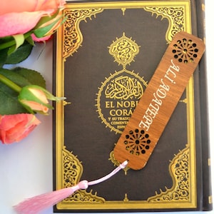 以下が含まれることがあります： ピンクのタッセルが付いた木製のブックマークで、スペイン語で「エル・ノブレ・コラン」と書かれています。ブックマークは、金色のアクセントとアラビア文字が書かれた茶色の本の上に置かれています。