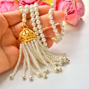 Può includere: Una collana di perle con un pendente e nappa in filigrana dorata. La collana è composta da più fili di perle bianche, con un cappuccio dorato decorativo e fili di perle pendenti. Rose rosa sullo sfondo.