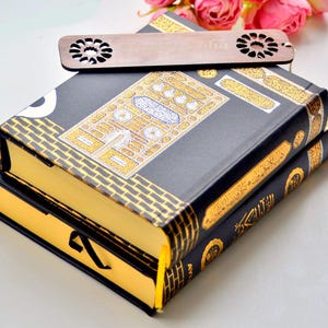 Könnte beinhalten: Zwei schwarze Bücher mit goldenen und weißen dekorativen Mustern und goldfarbenen Seiten sind gestapelt. Ein hölzerner Lesezeichen mit floralem Design liegt obenauf. Rosa Rosen sind im Hintergrund.