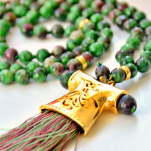 May include: A necklace with green and maroon beads. The necklace has a gold-coloured pendant with intricate cut-out designs and a tassel with green and maroon threads. The beads are round and strung together.