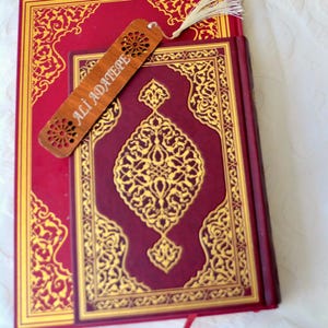 Peut inclure: Un livre rouge avec des motifs dorés et un marque-page en bois. Le marque-page porte le nom "ALI ADATEPE" et présente des découpes décoratives. Le livre a une bordure dorée et un motif doré central.