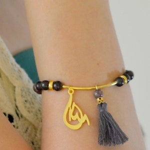 Può includere: Un braccialetto dorato con perline viola scuro, un ciondolo dorato con calligrafia araba e un fiocco grigio.