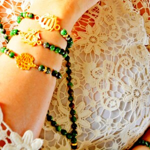 May include: A beaded bracelet and necklace set with green and brown beads and gold-coloured charms. The jewellery is displayed on an arm, with a cream lace top in the background. The charms have Arabic calligraphy.