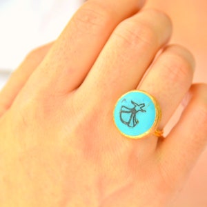 Può includere: Un anello color turchese con una montatura in oro presenta una silhouette nera di un derviscio roteante.