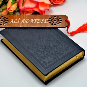 Könnte beinhalten: Ein Holzlesezeichen mit dem eingravierten Namen "ALI ADATEPE", verziert mit einem floralen Design und einem orangefarbenen Quastenanhänger. Das Lesezeichen liegt auf einem dunkelblauen Buch mit goldfarbenen Seiten.