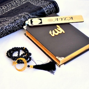 Op de afbeelding: Een zwart boek met goudkleurige randen en een gouden Arabisch schriftteken op de kaft, vergezeld van een houten bladwijzer gegraveerd met een hart en de naam AYLA, en een zwarte gebedsketting met een gouden halve maan hanger.