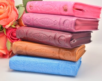 May include: A stack of five book-shaped zippered pouches in various colors: pink, purple, burgundy, tan, and blue. Each pouch has a textured, embossed design and a zipper closure. A bouquet of pink and red roses is in the background.