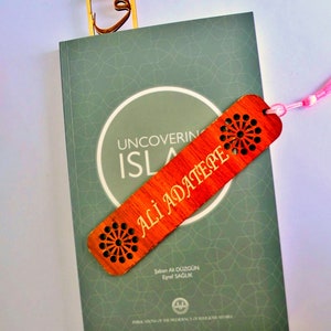 May include: A book titled "Uncovering Islam" with an orange bookmark featuring the name "Ali Adatepe" and a floral pattern. A wooden bookmark with a gold design is also visible, along with a tassel.