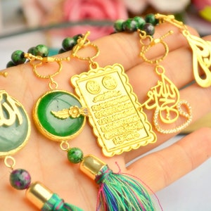 May include: A close-up of a gold-colored prayer bead necklace with green and red beads. The necklace features several gold pendants with Arabic calligraphy and decorative elements, including a green stone pendant and a tassel.